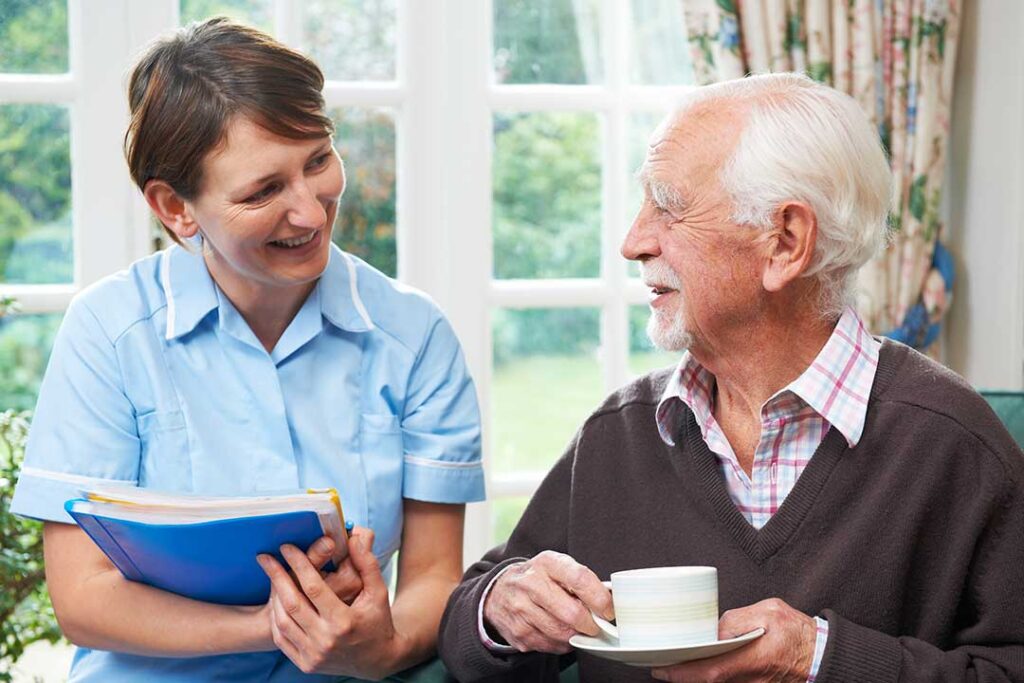 An elderly person sits in a cozy living room chair, holding hands with a visiting family member.